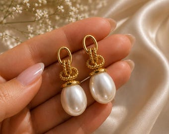 Op de afbeelding: Een paar oorbellen met glanzende witte ovale parels die aan goudkleurige metalen schakels hangen. Het ontwerp bevat een getextureerde, gedraaide, touwachtige gouden lus die de parel verbindt met de bovenste ovale schakel.