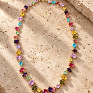 Pu&ograve; includere: Una collana a catena color oro caratterizzata da una serie di pietre preziose sfaccettate multicolori in varie forme, tra cui tagli ovali, rettangolari e a goccia. Le pietre mostrano una vivace tavolozza di tonalit&agrave; rosa, viola, gialle, blu e verdi.