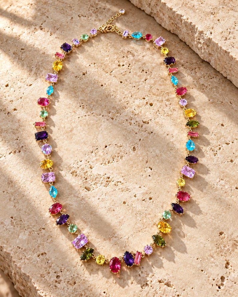 Pu&ograve; includere: Una collana a catena color oro caratterizzata da una serie di pietre preziose sfaccettate multicolori in varie forme, tra cui tagli ovali, rettangolari e a goccia. Le pietre mostrano una vivace tavolozza di tonalit&agrave; rosa, viola, gialle, blu e verdi.