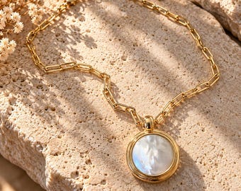 Op de afbeelding: Een goudkleurige ketting met een dikke, rechthoekige schakelketting en een grote, ronde hanger. De hanger heeft een gepolijst goudkleurig frame rondom een gladde, iriserende witte parelmoeren inleg in het midden.