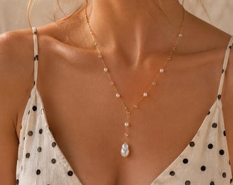 Op de afbeelding: Een gouden ketting met kleine witte parels die over de lengte zijn verdeeld en een enkele grotere, onregelmatig gevormde witte parel als hanger in het midden. De ketting wordt gedragen bij een crèmekleurige top met een zwart stippenpatroon.