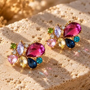 Pu&ograve; includere: Un paio di orecchini a lobo color oro con un gruppo di cristalli sfaccettati multicolori. Ogni orecchino presenta una grande pietra color magenta a forma di goccia, accentuata da cristalli pi&ugrave; piccoli rotondi, ovali e a pera nei colori rosa, giallo, blu, verde e trasparente.