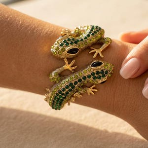 May include: A gold-toned cuff bracelet featuring two lizard figures facing each other. The lizards are encrusted with numerous small, sparkling green and yellow crystals, with dark, elongated eyes and textured, detailed legs and feet.