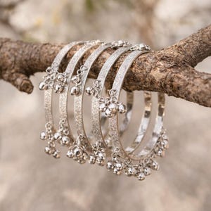 May include: Six silver bangles with dangling bell charms are displayed on a brown tree branch. The bangles have a textured, glittery appearance. The charms are small, round, and clustered together.