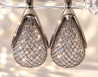 Op de afbeelding: Een paar druppelvormige oorbellen met een donker metalen frame dat een gaasrooster omsluit. Binnenin het gaas vangen talloze heldere, gefacetteerde kristallen het licht op, wat zorgt voor een sprankelend effect.