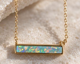 Op de afbeelding: Een goudkleurige ketting met een horizontale rechthoekige staafhanger ingelegd met een iriserende opaalsteen met flitsen van groen, blauw en oranje, hangend aan een fijne bijpassende goudkleurige schakelketting.