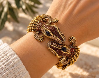 Op de afbeelding: Een goudkleurige armband met twee alligatorkoppen die naar elkaar toe zijn gericht. De lichamen van de alligators zijn ingelegd met kleine, glinsterende rode en amberkleurige kristallen, met donkere, opvallende ogen in elke kop.
