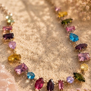 May include: A gold-tone necklace featuring a series of multi-coloured, faceted gemstones in various shapes including marquise, pear, and rectangular cuts. The stones display a vibrant palette of pink, purple, blue, yellow, and green hues.