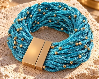 Op de afbeelding: Een meerstrengs armband bestaande uit gevlochten metallic turquoise draad, verweven met kleine, gepolijste goudkleurige kraaltjes. De armband wordt gesloten met een rechthoekige, gladde goudkleurige magnetische sluiting.