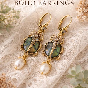 Puede incluir: Pendientes bohemios dorados con diseño de escarabajo, con detalles iridiscentes verdes y azules, pequeños cristales y una perla colgante. Los pendientes se muestran sobre una superficie de encaje blanco. Texto: Gift for Sister or Friend, BOHO EARRINGS.