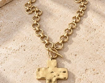 Op de afbeelding: Een goudkleurige ketting met een dikke schakelketting van ronde ringen en een grote kruishanger met een gehamerde textuur. De hanger is aan de ketting bevestigd met een staafsluiting, wat zorgt voor een opvallend en robuust sieraad.