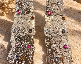 Op de afbeelding: Een paar zilverkleurige oorbellen met een gelaagd, getextureerd metalen ontwerp. Elke oorbel bestaat uit drie overlappende, onregelmatige rechthoekige plaatjes met een gehamerde afwerking, geaccentueerd met kleine, veelkleurige kristallen.
