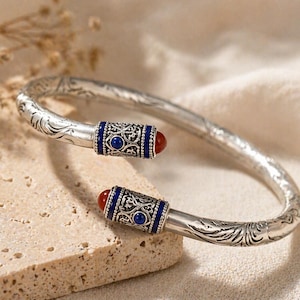 May include: A silver cuff bracelet featuring an intricate engraved floral pattern along the band. The open ends are capped with ornate silver cylinders, each inlaid with a deep blue lapis lazuli stone and a rounded, polished red carnelian bead.
