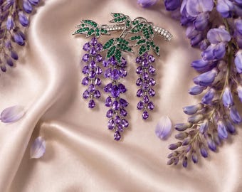 Op de afbeelding: Een zilverkleurige broche in de vorm van een blauweregentak, met drie hangende trossen van paarse druppelvormige stenen, groene bladvormige stenen en kleine heldere pavé-steentjes langs de steel, liggend op een gladde, lichtgekleurde stof.