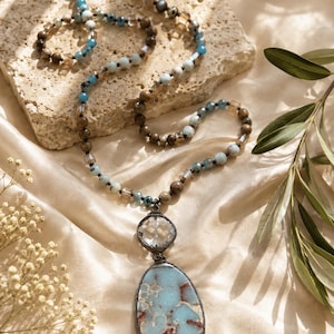 Può includere: Una lunga collana di perline con un mix di perline marroni, azzurre e trasparenti. Il ciondolo è composto da una pietra rotonda sfaccettata trasparente sopra una pietra più grande, irregolare e marmorizzata azzurra e marrone, in cornici scure.