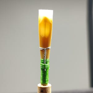 May include: A close-up of a reed instrument's reed. The reed is a light brown color with a white tip. It is wrapped with green thread and secured with silver wire. The reed is attached to a cork base.