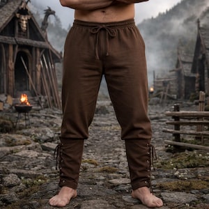 Op de afbeelding: Bruine broek met trekkoord en veterdetails bij de enkels. De broek heeft een relaxte pasvorm en is gemaakt van een zacht materiaal. De achtergrond toont een rustiek dorpsgezicht met houten structuren en een vuurplaats.