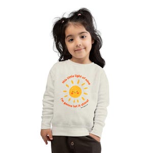 May include: A light beige sweatshirt featuring a smiling sun graphic with the text "this little light of mine, I'm gonna let it shine!" in orange. The sweatshirt is worn by a young child.