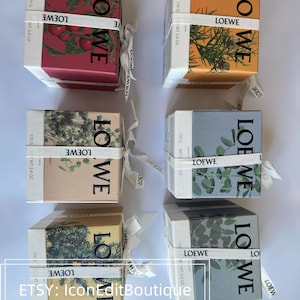 May include: Six square Loewe candle boxes arranged in two columns. Each box features a botanical illustration, a white ribbon with the Loewe logo, and text indicating a net weight of 170 grams or 5.8 ounces. The boxes are red, orange, beige, and blue.