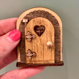 May include: A miniature wooden fairy door with a heart-shaped window. The door has a light brown finish with darker brown trim and hinges. A small knob and a decorative arch complete the design.