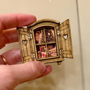 May include: A small, rectangular wooden window ornament with open shutters featuring heart-shaped cutouts. The center displays a printed scene of a cozy room with a decorated Christmas tree, a fireplace, wrapped gifts, and a plate of cookies.