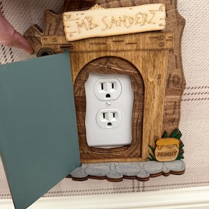 May include: A wooden wall outlet cover shaped like a tree trunk door with a blue hinged door panel. The cover features a sign reading MR SANDERZ and a small honey pot labeled HUNNY. The design surrounds a standard white dual electrical outlet.