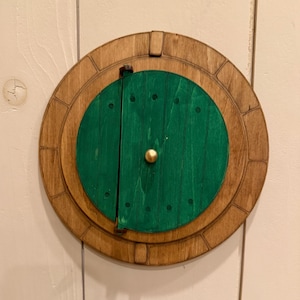 May include: A circular wooden wall decoration featuring a green door with vertical planks and a central gold-colored knob. The door is set within a light brown wooden frame that has a segmented, circular border design with dark etched lines.