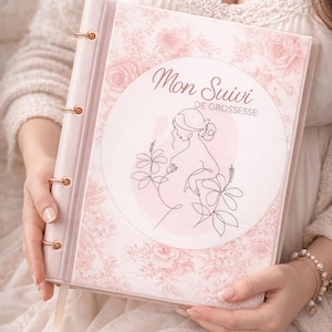 Könnte beinhalten: Ein rosa Schwangerschaftstagebuch mit goldenen Akzenten und dem französischen Text "Mon Suivi de Grossesse". Das Tagebuch hat ein Blumenmuster und eine Strichzeichnung einer schwangeren Person. Das Tagebuch ist geöffnet.