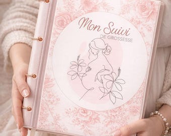 Op de afbeelding: Een roze zwangerschapsdagboek met gouden accenten en de Franse tekst "Mon Suivi de Grossesse". Het dagboek heeft een bloemenpatroon en een lijntekening van een zwangere persoon. Het dagboek is open.