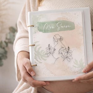 Peut inclure: Un classeur blanc avec des accents dorés, orné d'un motif aquarelle et du texte "Mon Suivi". La couverture présente un dessin au trait d'une personne enceinte et des éléments floraux. Le classeur est tenu devant une personne.