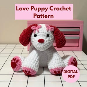 Puede incluir: Un cachorro de ganchillo blanco con orejas, nariz y almohadillas rojas. El cachorro tiene ojos negros y lazos rosas. La imagen incluye el texto "Love Puppy Crochet Pattern" y "DIGITAL PDF".