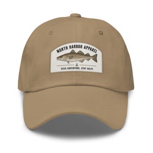 May include: A tan baseball cap featuring a rectangular patch with a fish illustration and the text "NORTH HARBOR APPAREL." Below the illustration, the text reads "SEEK ADVENTURE. STAY SALTY."