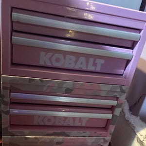 以下が含まれることがあります： ピンク色のKobaltツールチェスト。複数の引き出しがあり、引き出しにはシルバーのハンドルが付いています。Kobaltのロゴは白で印刷されています。下の引き出しには迷彩柄があります。