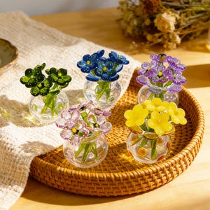 May include: Five small glass vases, each holding a different colored glass flower arrangement. The flowers are green, blue, purple, pink, and yellow. The vases sit on a woven tray, with a neutral-colored cloth and a lit candle in the background.