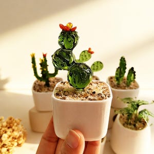 May include: A small, green glass cactus sculpture with a tiny orange flower on top, potted in a white square ceramic container filled with light-colored pebbles. The cactus features a rounded, segmented body and is held in a hand.