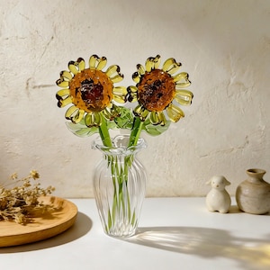 May include: Two glass sunflowers with yellow petals and brown centers are displayed in a clear, fluted glass vase. The stems are green. The vase sits on a white surface with a wooden plate and small ceramic figurines in the background.