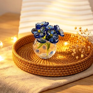 May include: A small, clear glass vase with blue glass flowers, set on a round, woven rattan tray. The flowers have a dark blue hue with yellow centers. The tray sits on a beige linen cloth, creating a natural, decorative display.