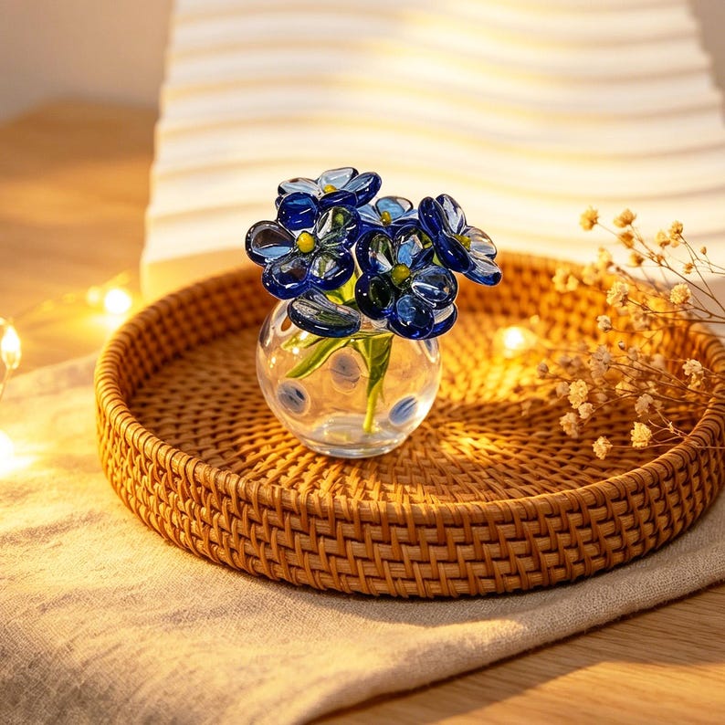 May include: A small, clear glass vase with blue glass flowers, set on a round, woven rattan tray. The flowers have a dark blue hue with yellow centers. The tray sits on a beige linen cloth, creating a natural, decorative display.