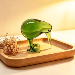 May include: A translucent green glass figurine shaped like a kiwi bird with a long, pointed beak and detailed feet. The bird is standing on a smooth, light-colored wooden tray next to a small cluster of dried, off-white baby's breath flowers.