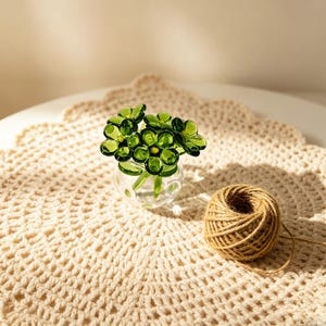May include: A small glass vase filled with green glass flowers. The vase sits on a cream-colored, crocheted doily. A ball of brown twine rests nearby, suggesting a craft or decorative theme.