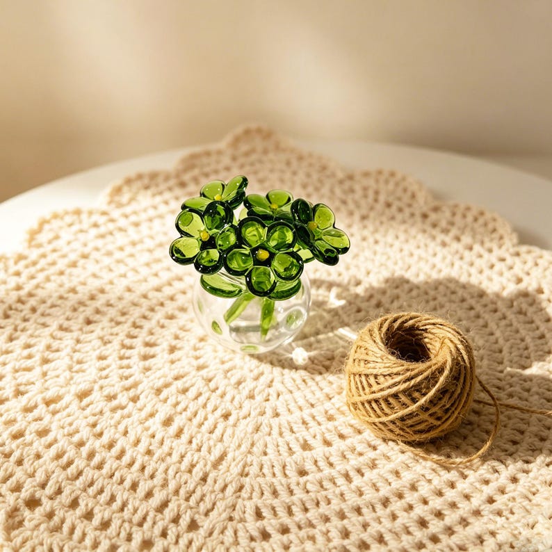 May include: A small glass vase filled with green glass flowers. The vase sits on a cream-colored, crocheted doily. A ball of brown twine rests nearby, suggesting a craft or decorative theme.