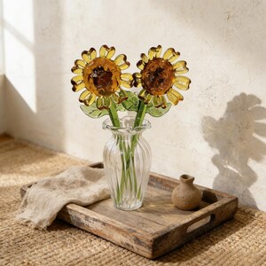 May include: Two glass sunflower stems in a clear, ribbed vase. The sunflowers have yellow petals and brown centers. The vase sits on a wooden tray with a small, brown ceramic vase and a beige linen cloth.