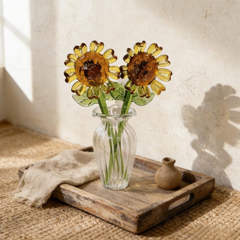 May include: Two glass sunflower stems in a clear, ribbed vase. The sunflowers have yellow petals and brown centers. The vase sits on a wooden tray with a small, brown ceramic vase and a beige linen cloth.