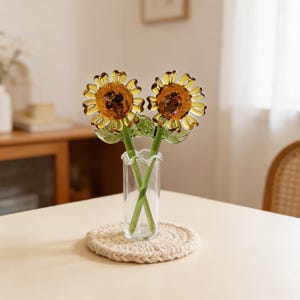 May include: Two glass sunflower stems in a clear vase with a ruffled top. The sunflowers have yellow petals and brown centers. The stems are green. The vase sits on a beige, round, woven coaster.