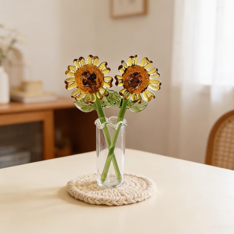 May include: Two glass sunflower stems in a clear vase with a ruffled top. The sunflowers have yellow petals and brown centers. The stems are green. The vase sits on a beige, round, woven coaster.