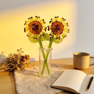 May include: Two glass sunflower sculptures in a clear vase. The sunflowers have yellow petals and brown centers. The stems are green. The vase is on a wooden table with a notebook, pen, and mug.
