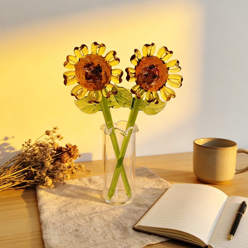 May include: Two glass sunflower sculptures in a clear vase. The sunflowers have yellow petals and brown centers. The stems are green. The vase is on a wooden table with a notebook, pen, and mug.
