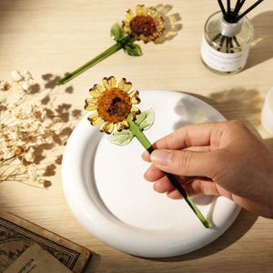 May include: Two glass sunflower ornaments with yellow petals and a brown center. The stems and leaves are green. One ornament is held over a white plate. The background includes a diffuser and dried flowers.