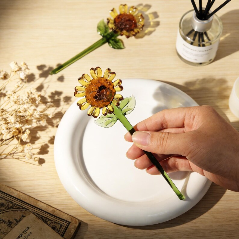 May include: Two glass sunflower ornaments with yellow petals and a brown center. The stems and leaves are green. One ornament is held over a white plate. The background includes a diffuser and dried flowers.