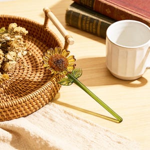 May include: A decorative glass sunflower with a yellow and brown flower head, green stem, and leaves. The sunflower is next to a woven basket, a white mug, and books. The sunflower is a unique home decor item.